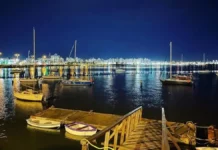 Veleiros ancorados à noite na Baía Sul, com luzes da cidade refletidas na água. Imagem ilustra a 1ª Regata Plínio Verani de Veleiros de Oceano, em São José.