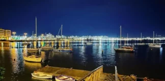 Veleiros ancorados à noite na Baía Sul, com luzes da cidade refletidas na água. Imagem ilustra a 1ª Regata Plínio Verani de Veleiros de Oceano, em São José.