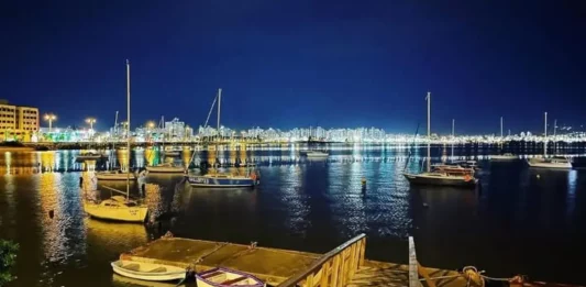 Veleiros ancorados à noite na Baía Sul, com luzes da cidade refletidas na água. Imagem ilustra a 1ª Regata Plínio Verani de Veleiros de Oceano, em São José.
