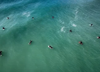 Florianópolis abre 307 vagas para atividades nas praias durante o verão Vista aérea de surfistas no mar, ilustrando as autorizações para aulas de surfe e outras atividades nas praias de Florianópolis durante o verão 2026/2027.