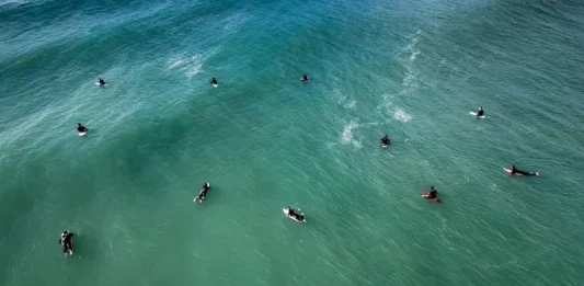 Florianópolis abre 307 vagas para atividades nas praias durante o verão Vista aérea de surfistas no mar, ilustrando as autorizações para aulas de surfe e outras atividades nas praias de Florianópolis durante o verão 2026/2027.