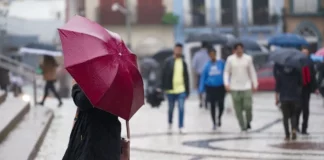 Pessoa segura guarda-chuva vermelho enquanto pedestres caminham sob chuva em uma área urbana de Santa Catarina.