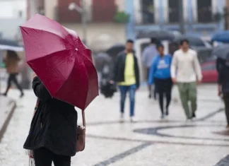 Pessoa segura guarda-chuva vermelho enquanto pedestres caminham sob chuva em uma área urbana de Santa Catarina.