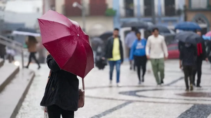 temporais_santa_catarina_1200x675 Pessoa segura guarda-chuva vermelho enquanto pedestres caminham sob chuva em uma área urbana de Santa Catarina.